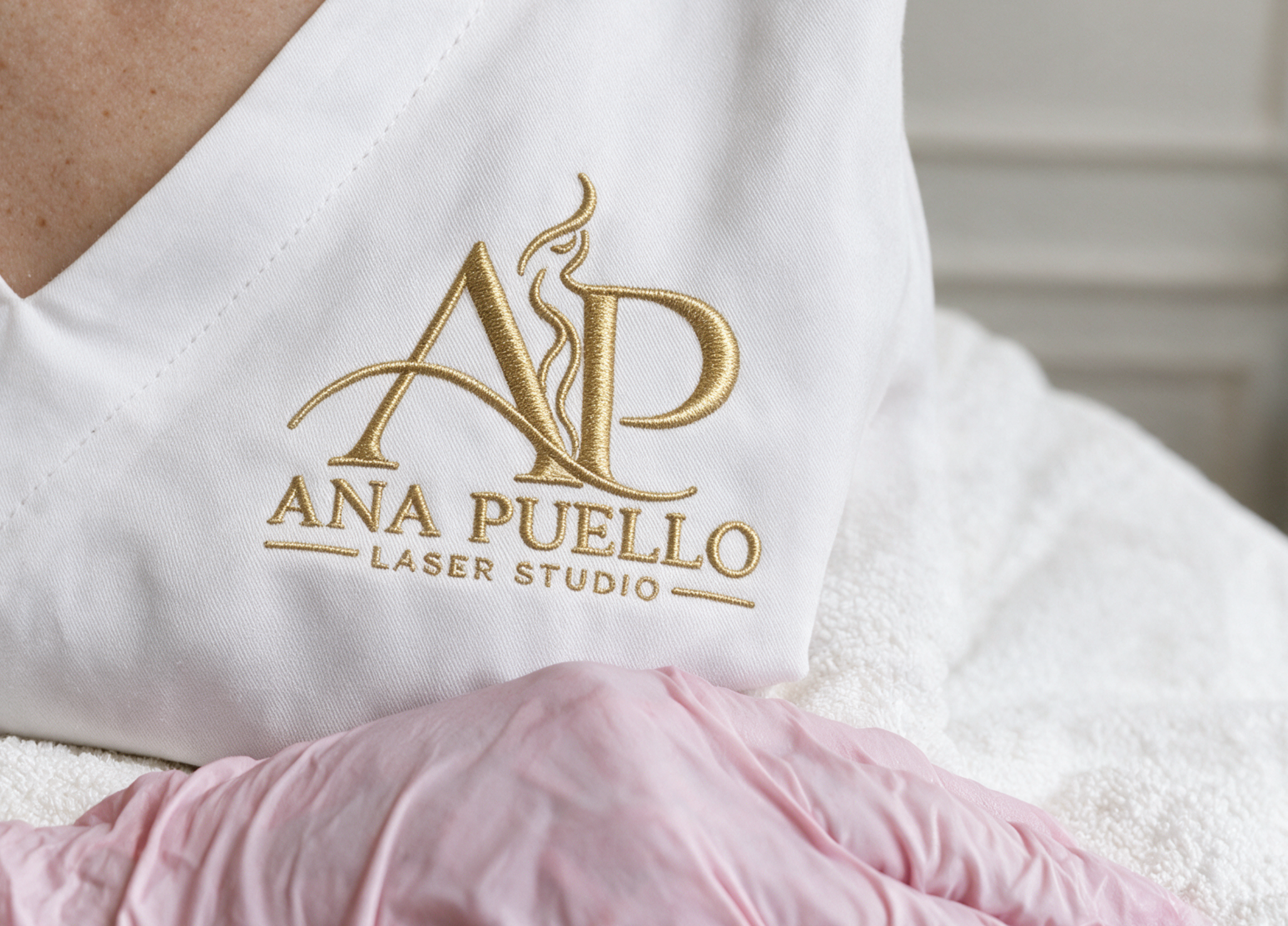 Logo dorado de Ana Puello Laser Studio bordado sobre uniforme blanco, junto a guantes rosados
